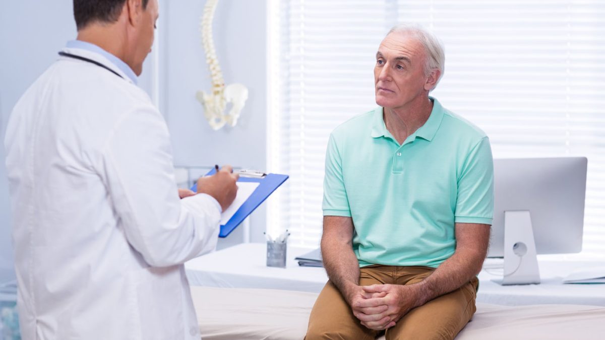 Older-man-on-a-medical-exam-table-listening-to-a-doctor-with-a-clipboard-in-a-well-lit-clinic-room